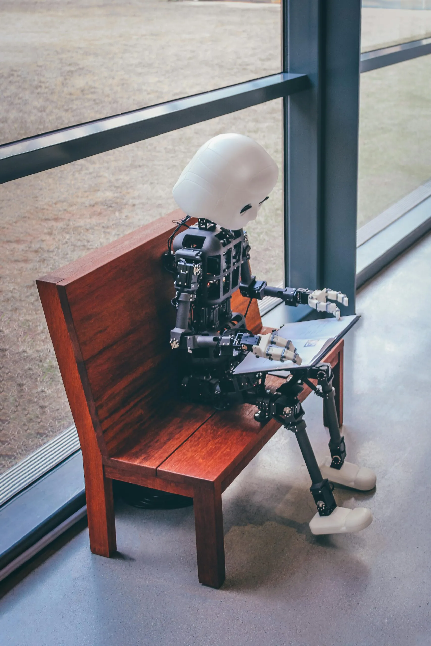 Bild på en robot sittandes på en bänk vid ett fönster, med en laptop i knät.